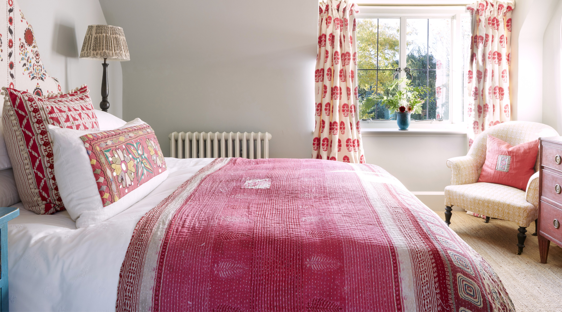 a comfy bed with white bedsheets and a pink throw, with embroidered cushions in a light and airy room